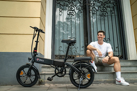 All-black lightweight DYU A1F PRO folding electric bike with dual-seat design and small urban tires, set on paved ground next to wrought iron glass doorway as young man rests casually on stone stairs