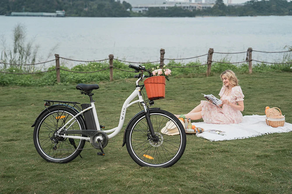 Lifestyle shot of white DYU C6 step-through city electric bike with flower basket on lakeside lawn, blonde woman relaxing on picnic blanket beside it.