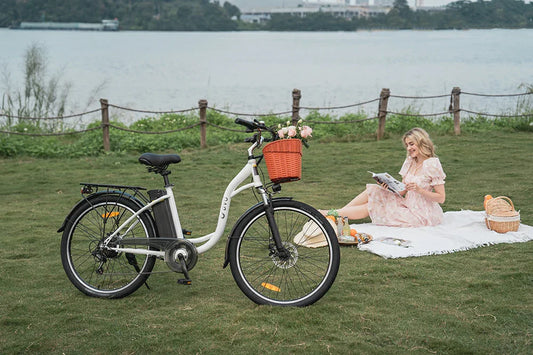 Lifestyle shot of white DYU C6 step-through city electric bike with flower basket on lakeside lawn, blonde woman relaxing on picnic blanket beside it.