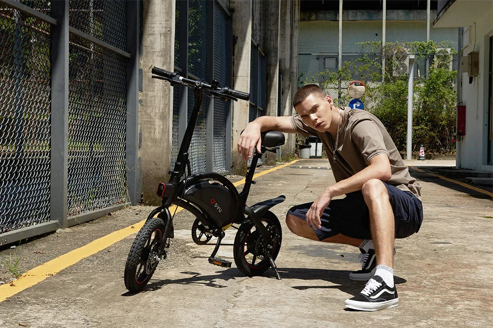 Mini-sized black DYU D3F lightweight foldable electric bicycle with red-rim tires, stylish male model squatting next to compact ebike on concrete industrial roadside